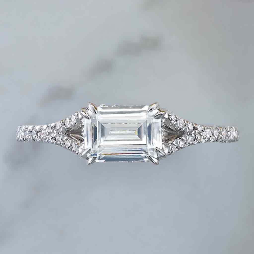 East-west French-set diamond halo engagement ring with emerald-cut center, editorial view