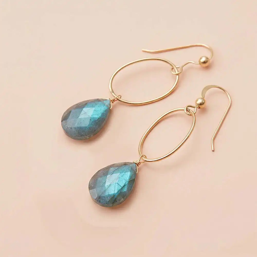 Faceted labradorite drop earrings in yellow gold fill, grey gemstone
teardrop briolette with blue-green flash, suspended from slim oval
gold link, blush background. all-groups