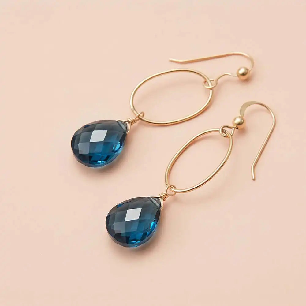 Faceted London blue topaz drop earrings in yellow gold fill, deep teal-blue gemstone teardrop briolette suspended from slim oval gold link, blush background. all-groups