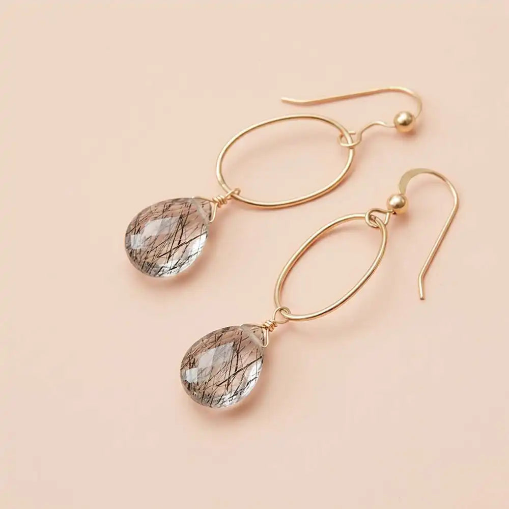 Faceted rutilated quartz drop earrings in yellow gold fill, clear gemstone teardrop briolette with golden needle inclusions, suspended from slim oval gold link, blush background. all-groups