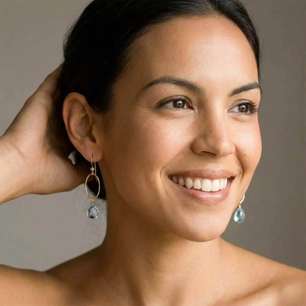 Woman wearing small faceted Swiss blue topaz hoop earrings in yellow gold fill,
bright medium-blue gemstone with clear vivid flash, lifestyle portrait. all-groups