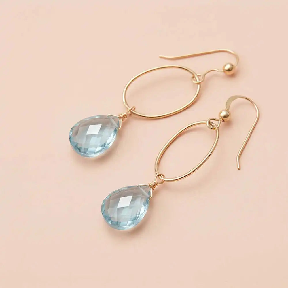 Faceted Swiss blue topaz drop earrings in yellow gold fill, bright medium-blue gemstone teardrop briolette suspended from slim oval gold link, blush background. all-groups